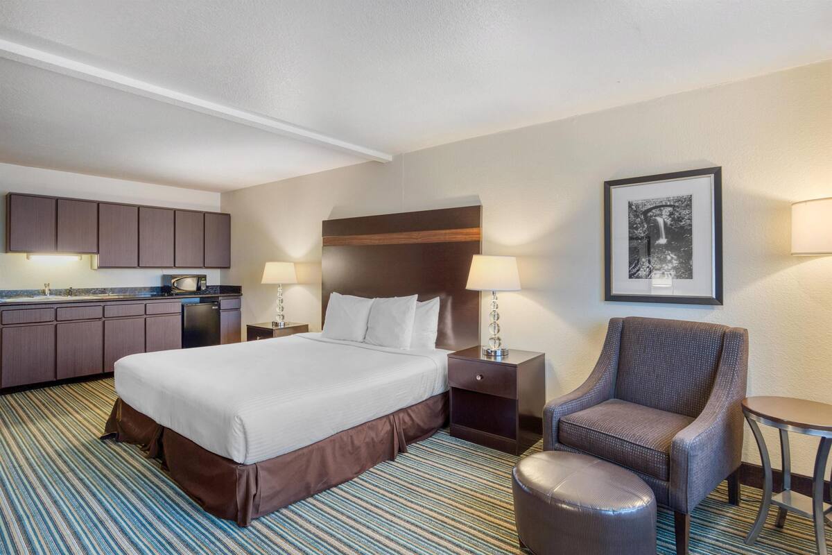 A hotel room with a large bed, a chair, a small table, a nightstand, a lamp, a framed picture on the wall, and a kitchenette with cabinets and a countertop.