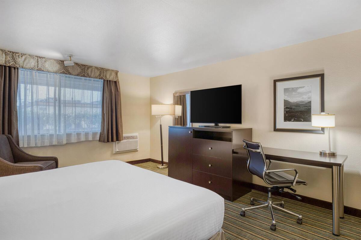 A hotel room with a bed, a desk with a computer, a chair, a television, and a window with curtains.