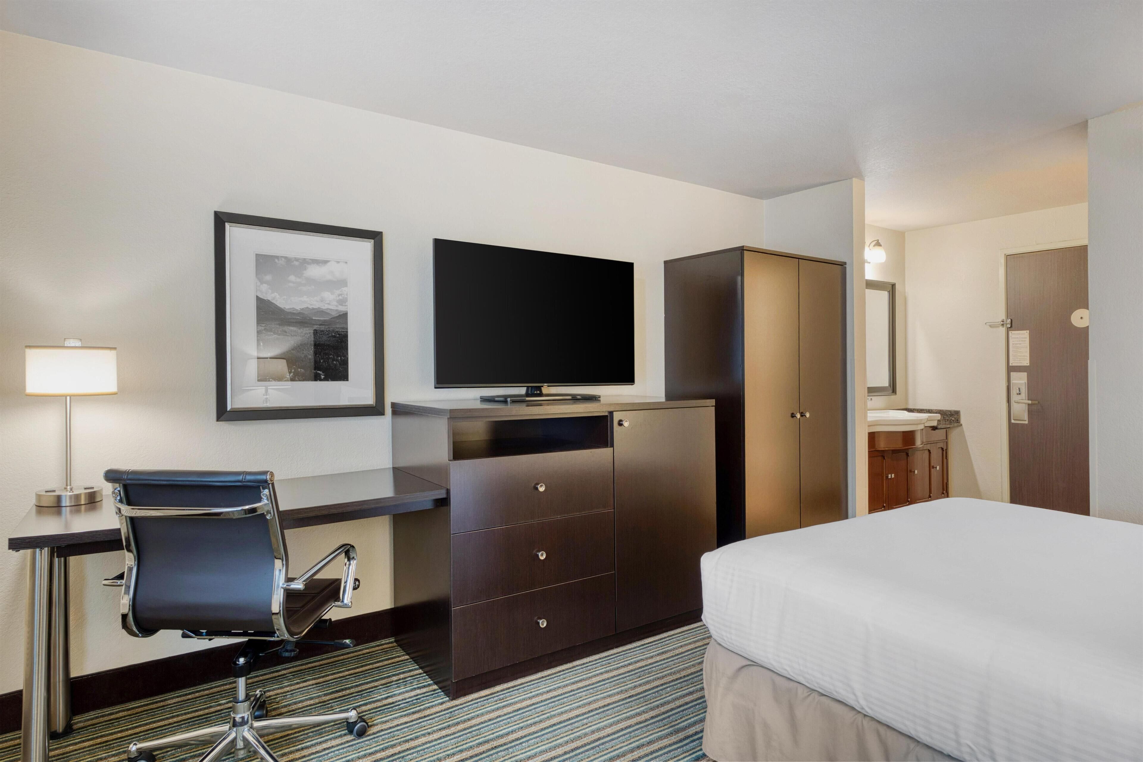 A hotel room with a bed, a desk with a chair, a television, a wardrobe, and a mirror.
