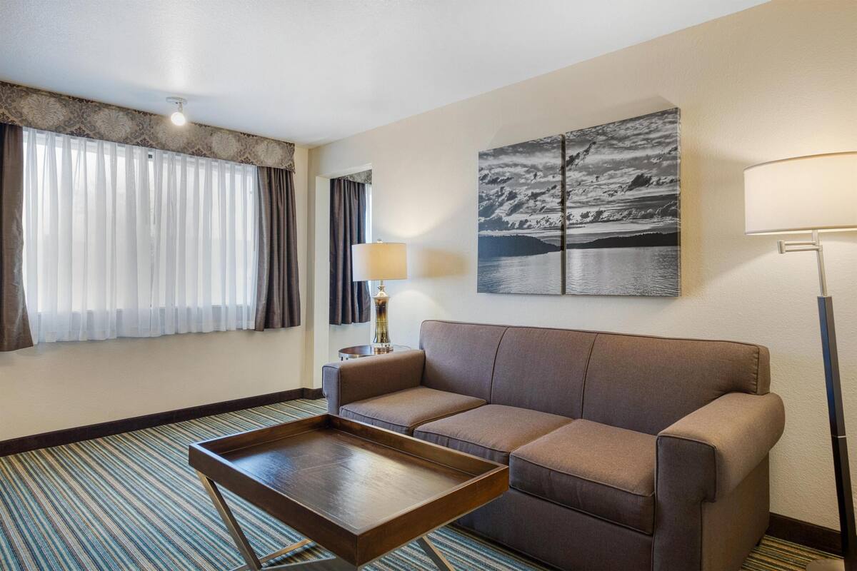 A hotel room with a sofa, a small table, a lamp, and a large landscape painting on the wall.