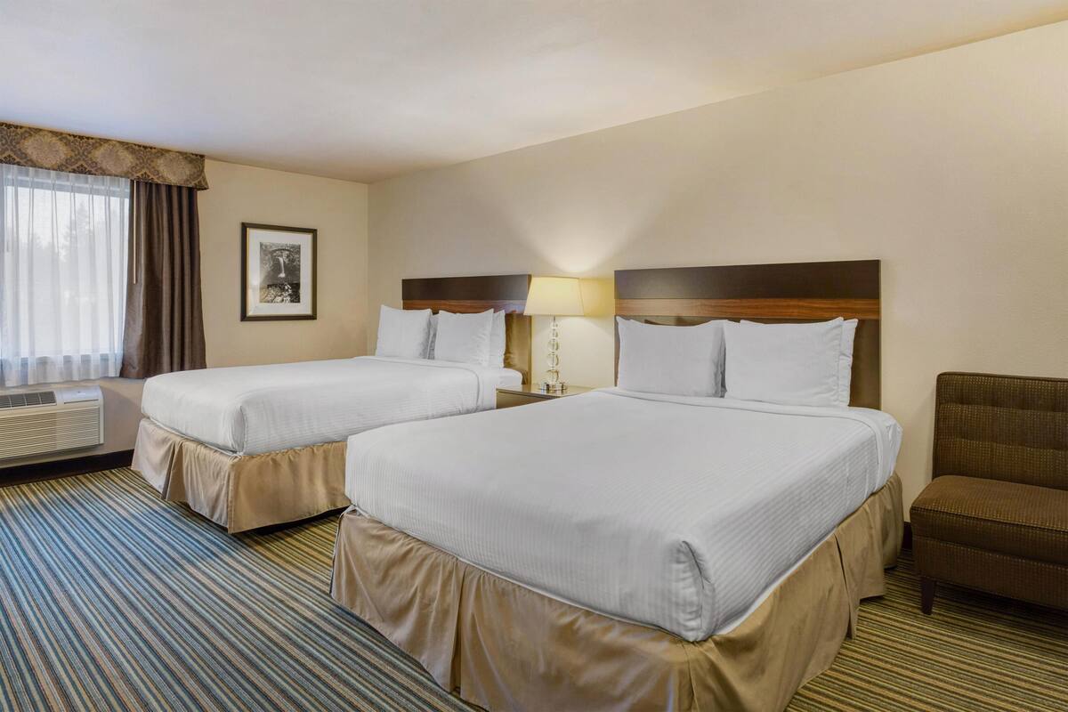 A hotel room with two beds, a chair, a lamp, and a framed picture on the wall.