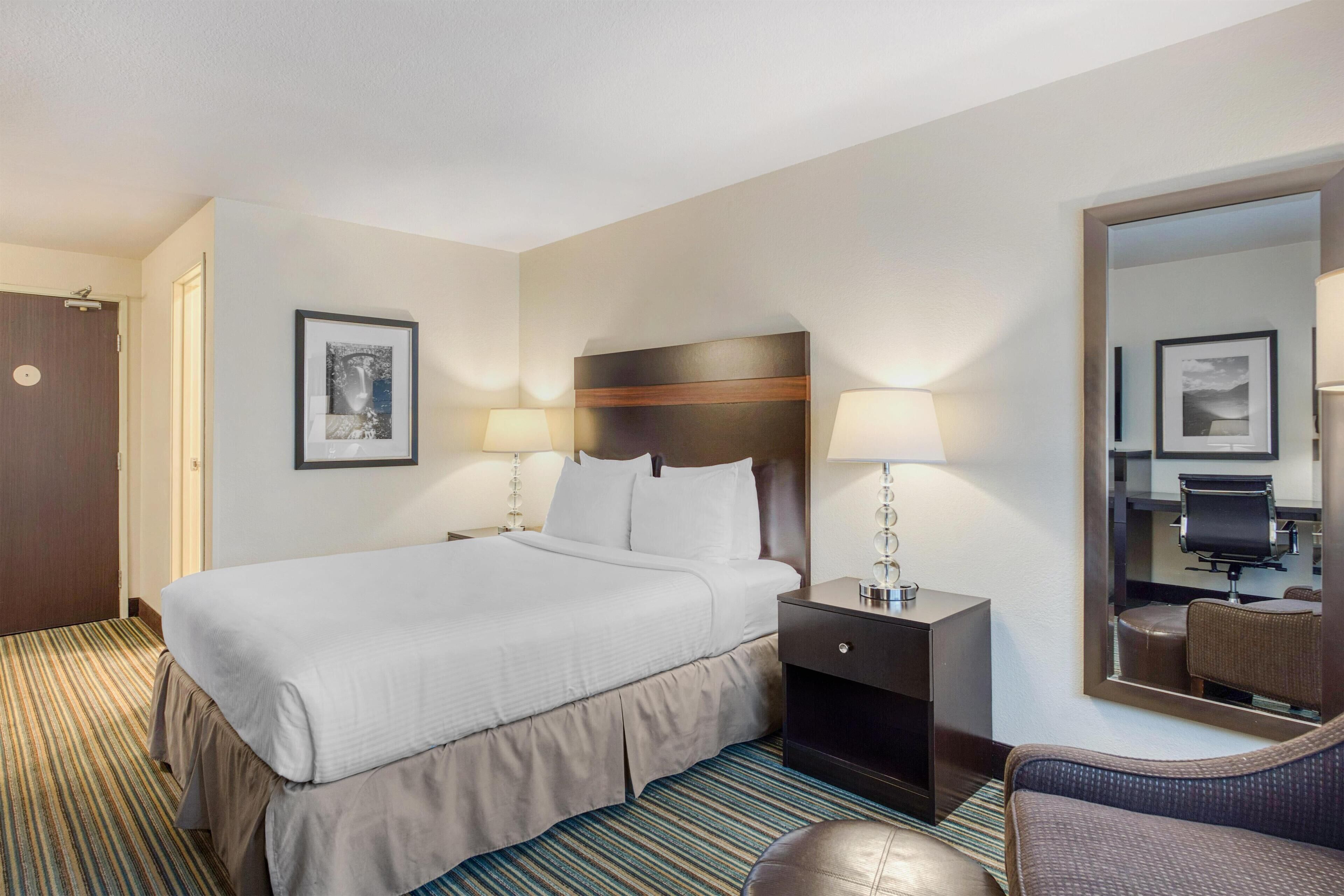 A hotel room with a large bed, two bedside tables with lamps, a chair, a mirror, and a door.