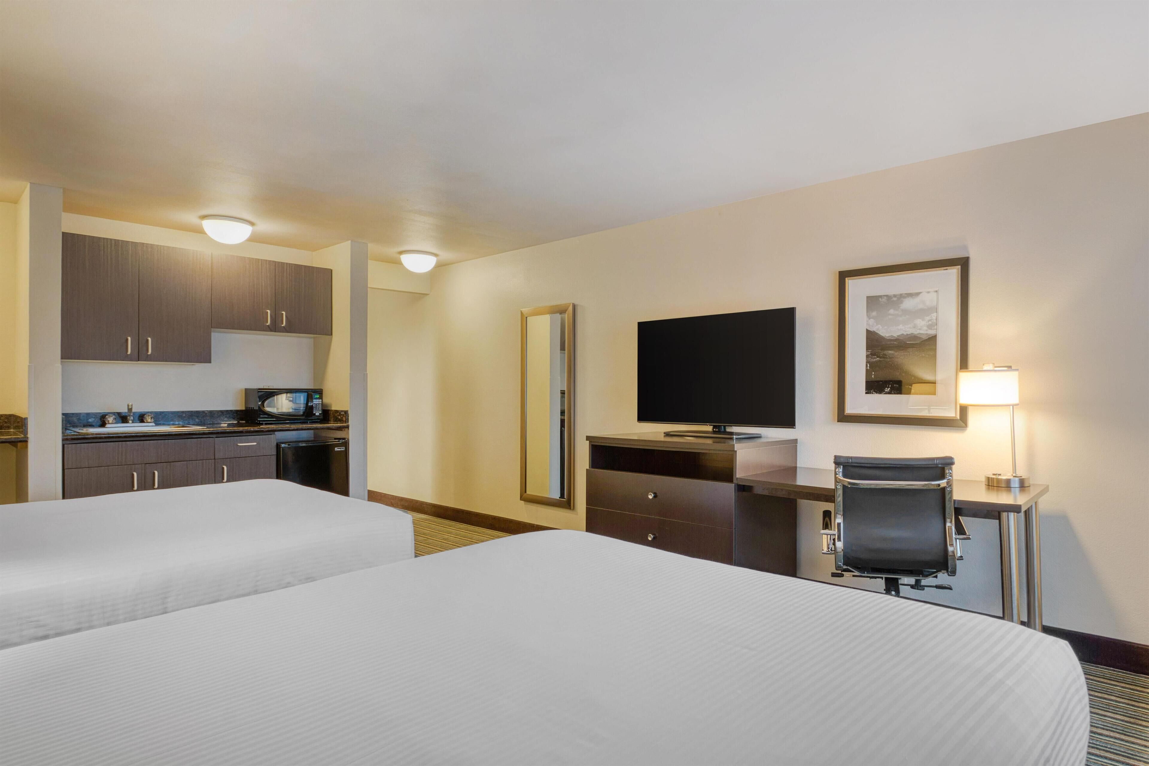 A hotel room with two beds, a desk with a chair, a television, a mirror, and a kitchenette.