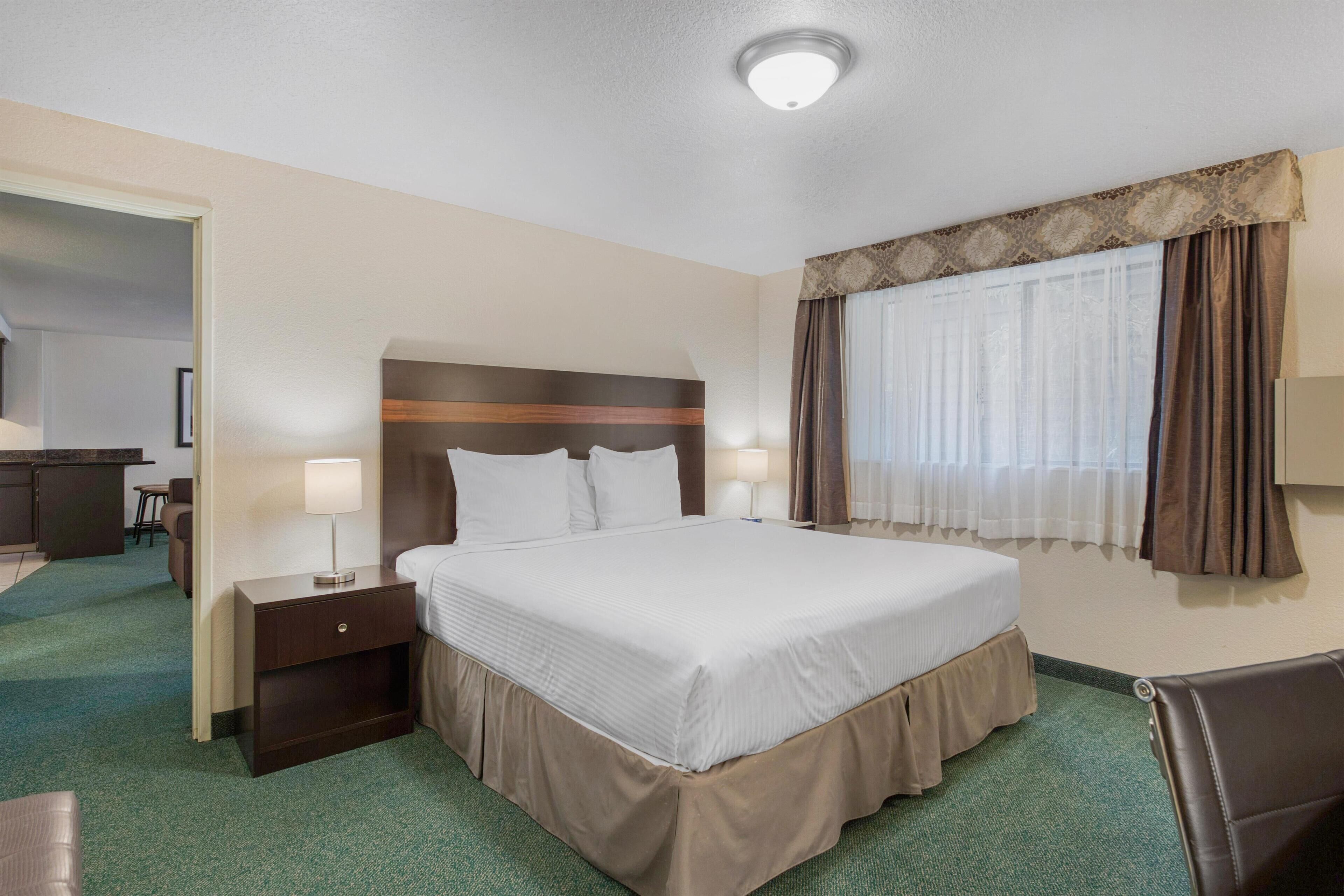 Ein Hotelzimmer mit einem großen Bett, einem Nachttisch, einem Schreibtisch und einem Fenster mit Vorhängen.