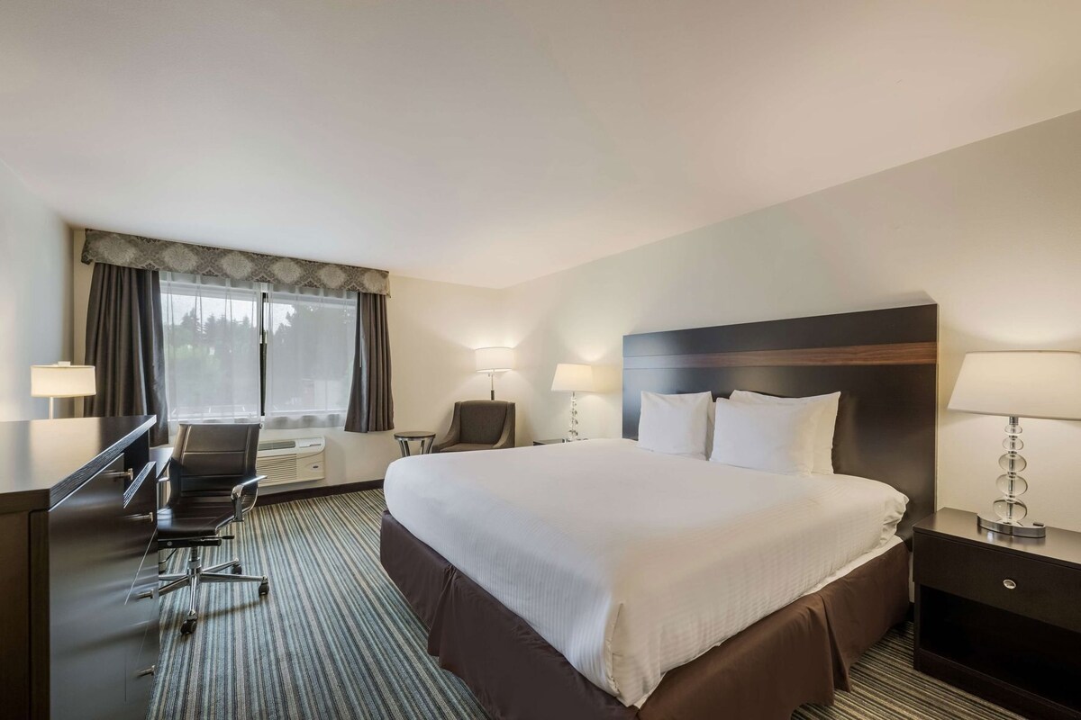 A hotel room with a large bed, a desk, a chair, a small table, and a lamp.