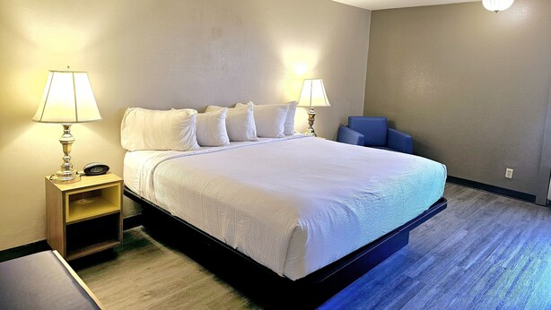 Una cama bien tendida en una habitación de hotel, con una mesita de noche de madera, dos lámparas de noche y una silla azul.