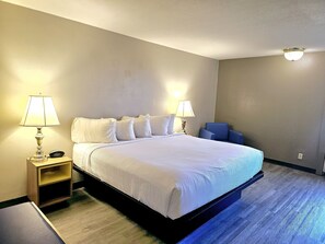 Una cama bien tendida en una habitación de hotel, con una mesita de noche de madera, dos lámparas de noche y una silla azul.
