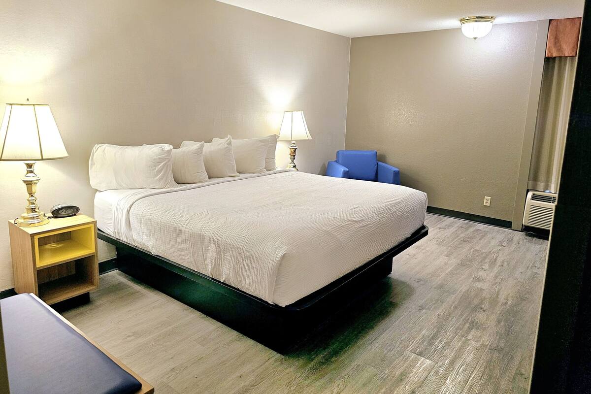 A hotel room with a bed, bedside lamps, a chair, a small table, and an air conditioning unit.