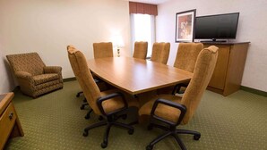 Una sala de conferencias con una gran mesa de madera, seis sillas tapizadas, un televisor sobre un mueble bajo y una ventana con cortinas.
