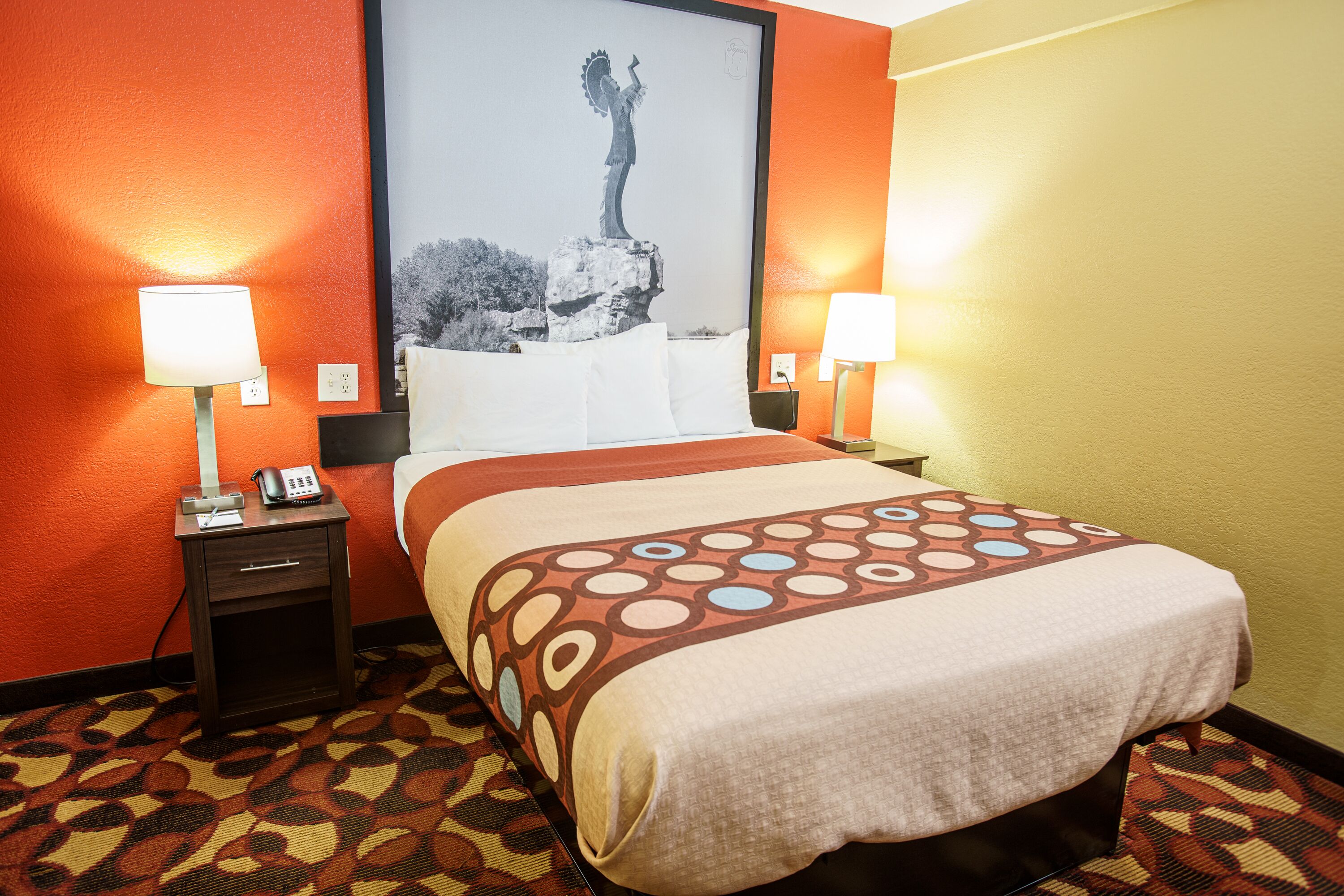 Une chambre d’hôtel avec un lit, deux tables de chevet avec des lampes, une moquette à motifs et un grand tableau encadré au mur.