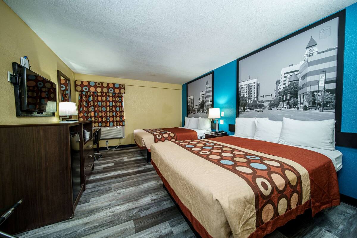 A hotel room with two beds, a desk, a chair, a mirror, and a large window with a cityscape view.