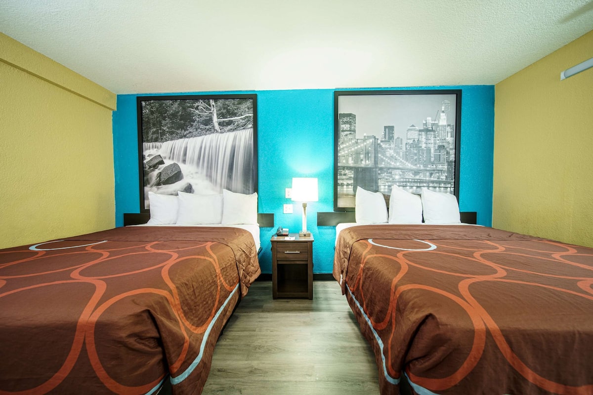 A hotel room with two beds, a nightstand with a lamp, and two large framed pictures on the walls.