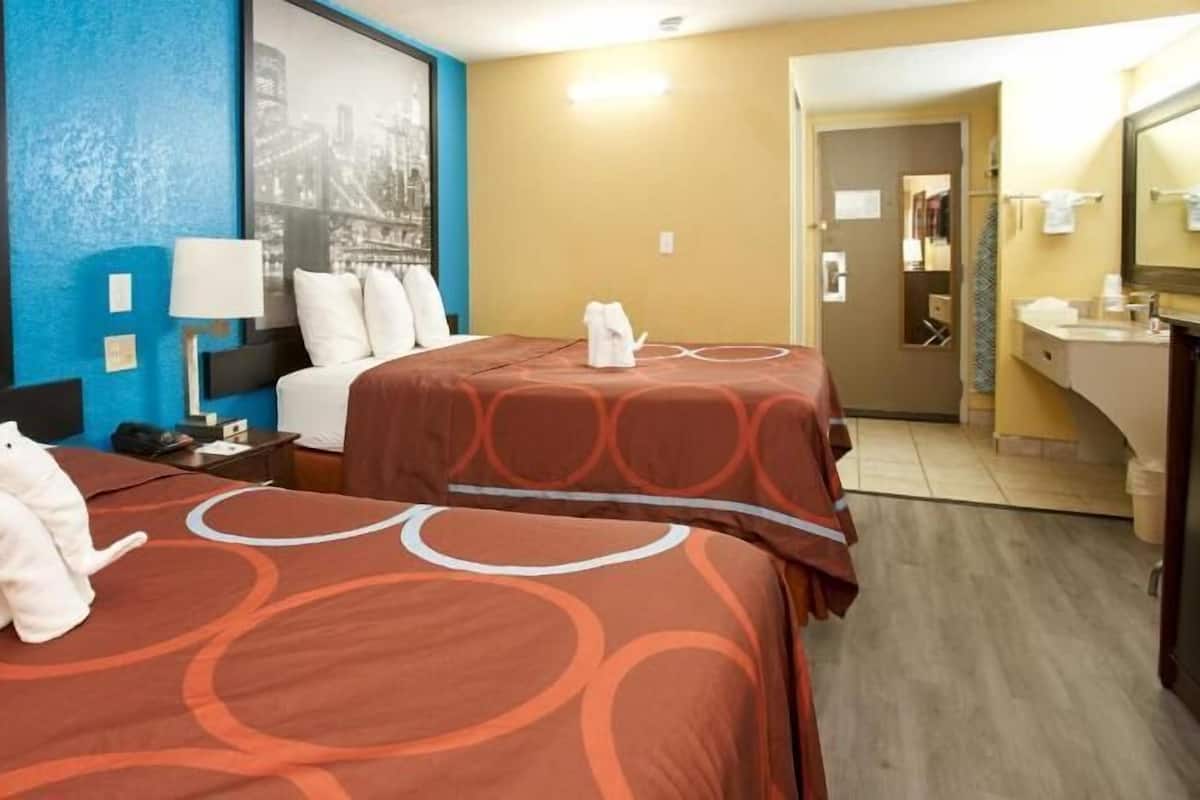 A hotel room with two beds, a large window with a cityscape view, a bathroom with a sink and mirror, and a hallway leading to another room.