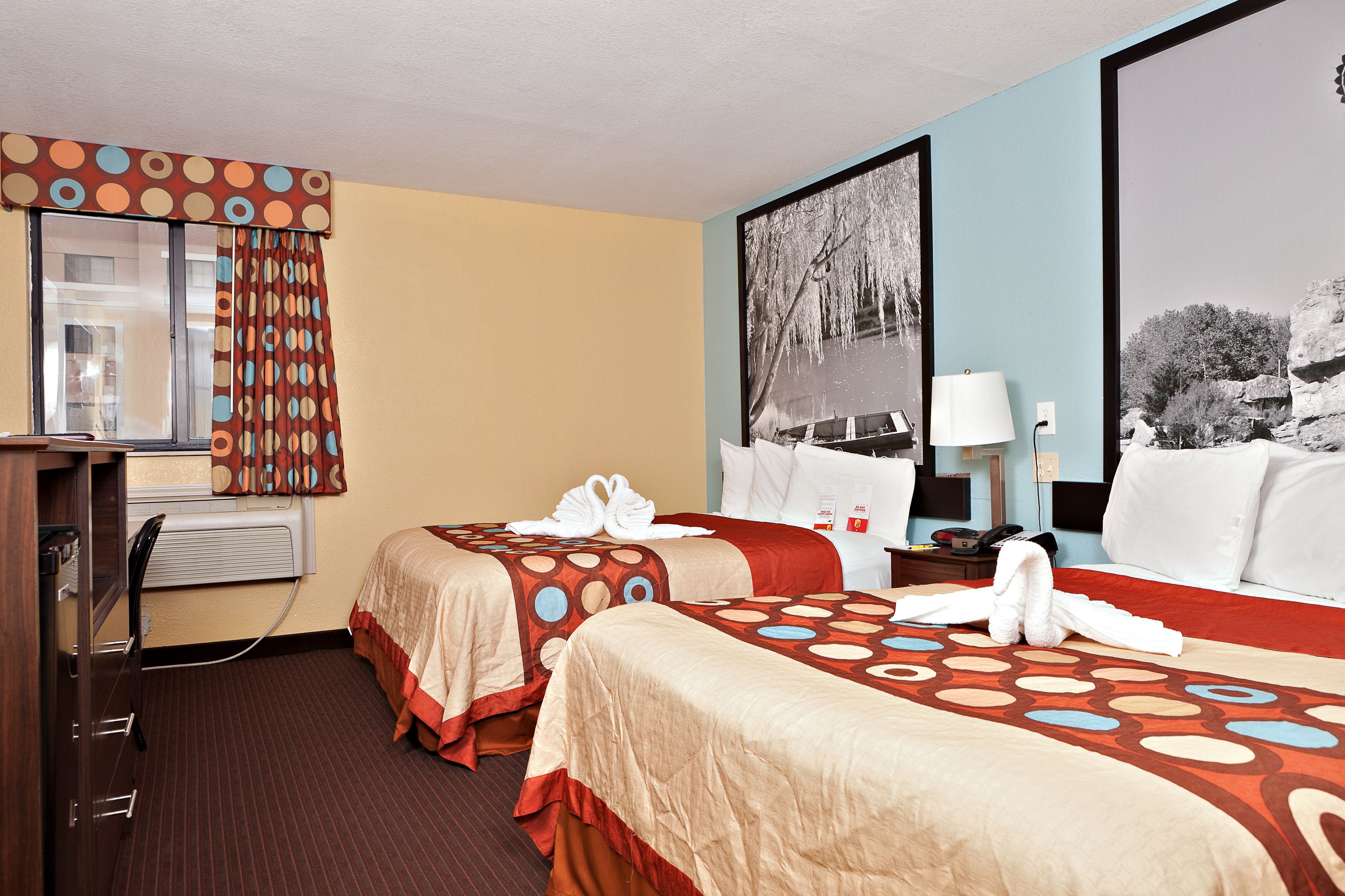 Una camera d'albergo con due letti, una scrivania, una sedia, una finestra con tende, una lampada e un quadro appeso al muro.