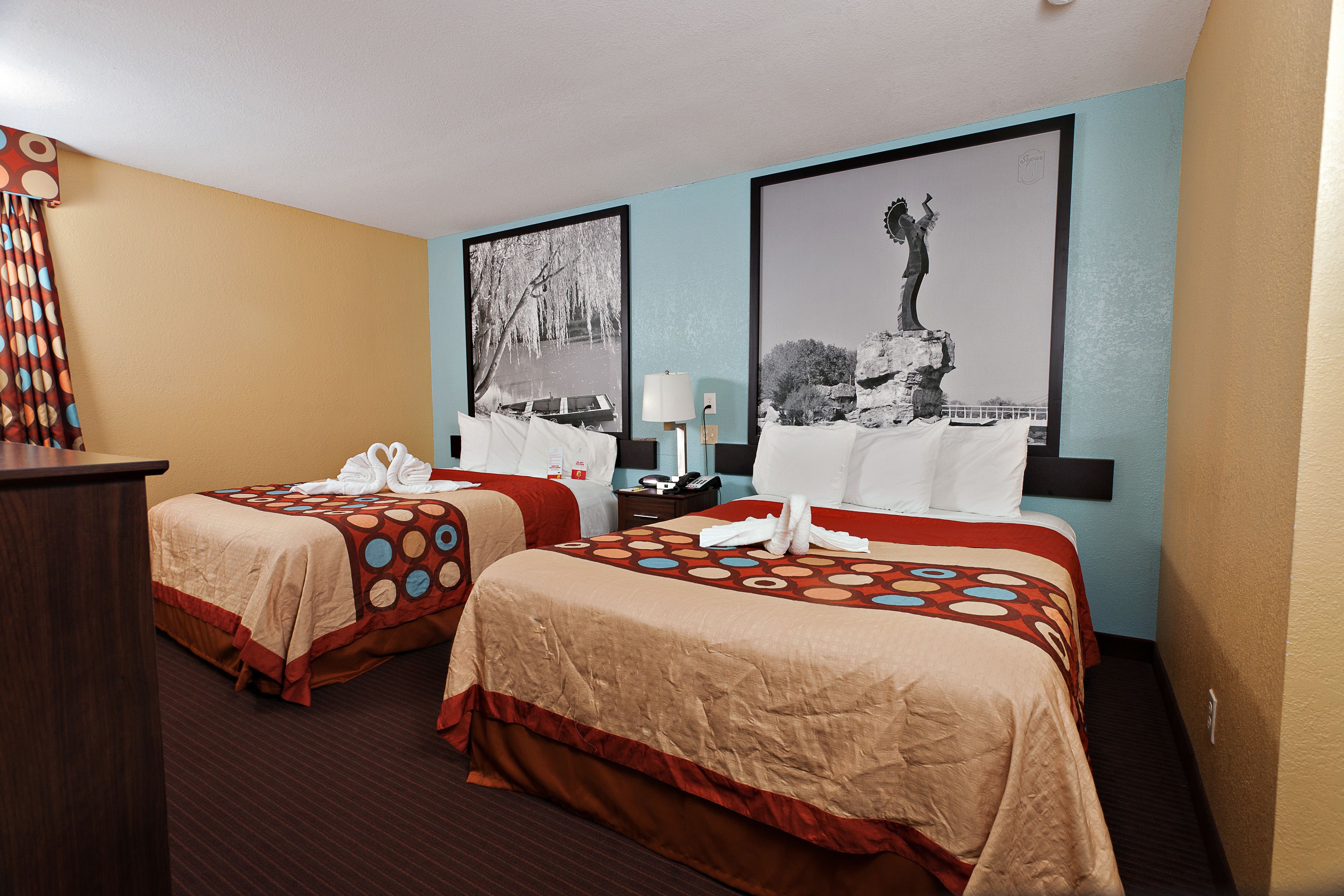 Ein Hotelzimmer mit zwei Betten, einem Schreibtisch und einem Nachttisch. An der Wand hängen zwei gerahmte Bilder.