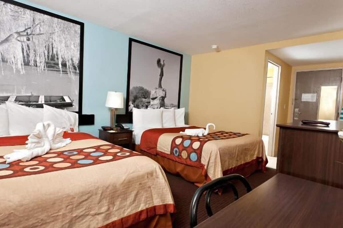 A hotel room with two beds, a desk, a chair, a desk lamp, a potted plant, and a painting on the wall.