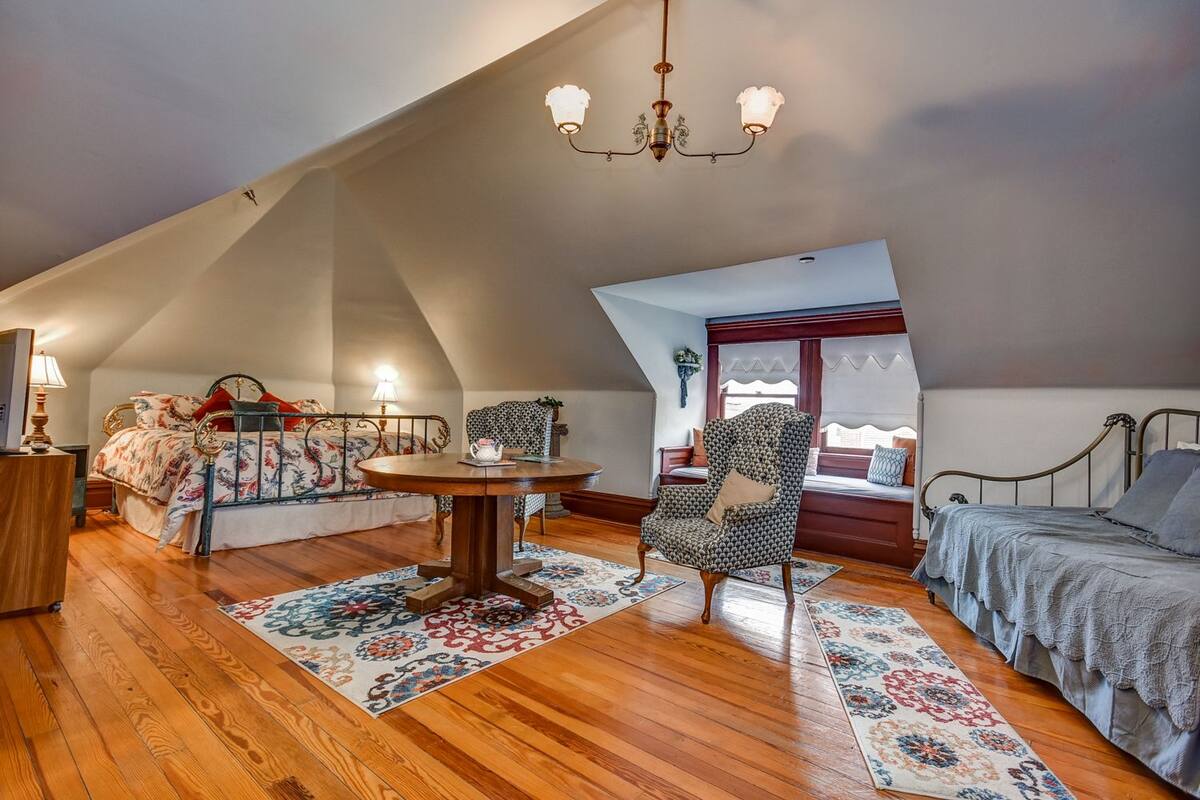 A cozy attic bedroom with a sloped ceiling, a round wooden table, a patterned armchair, and a bed with a patterned bedspread.