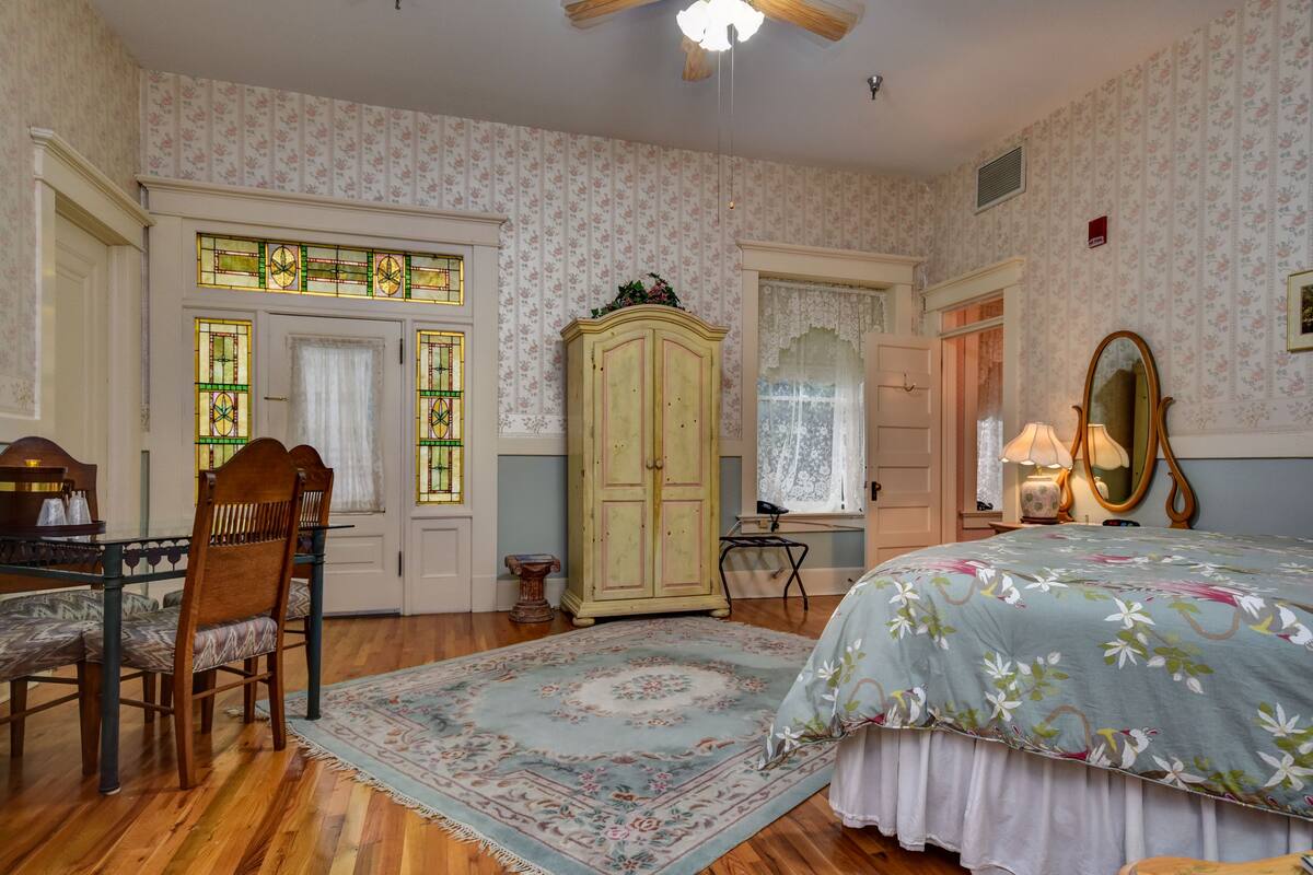 A bedroom with floral wallpaper, a wooden floor, a bed with a floral bedspread, a wooden chair, a cabinet, and a mirror.
