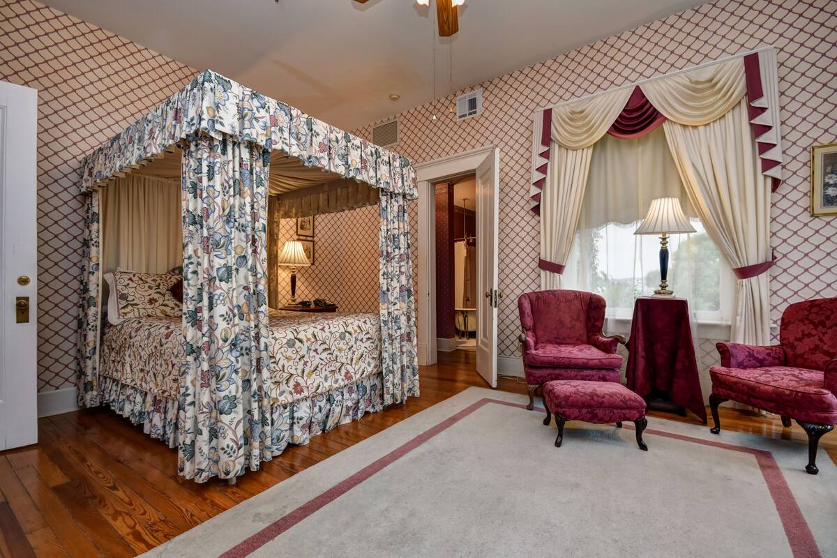 A bedroom with a canopy bed, two red armchairs, a table, and patterned wallpaper.