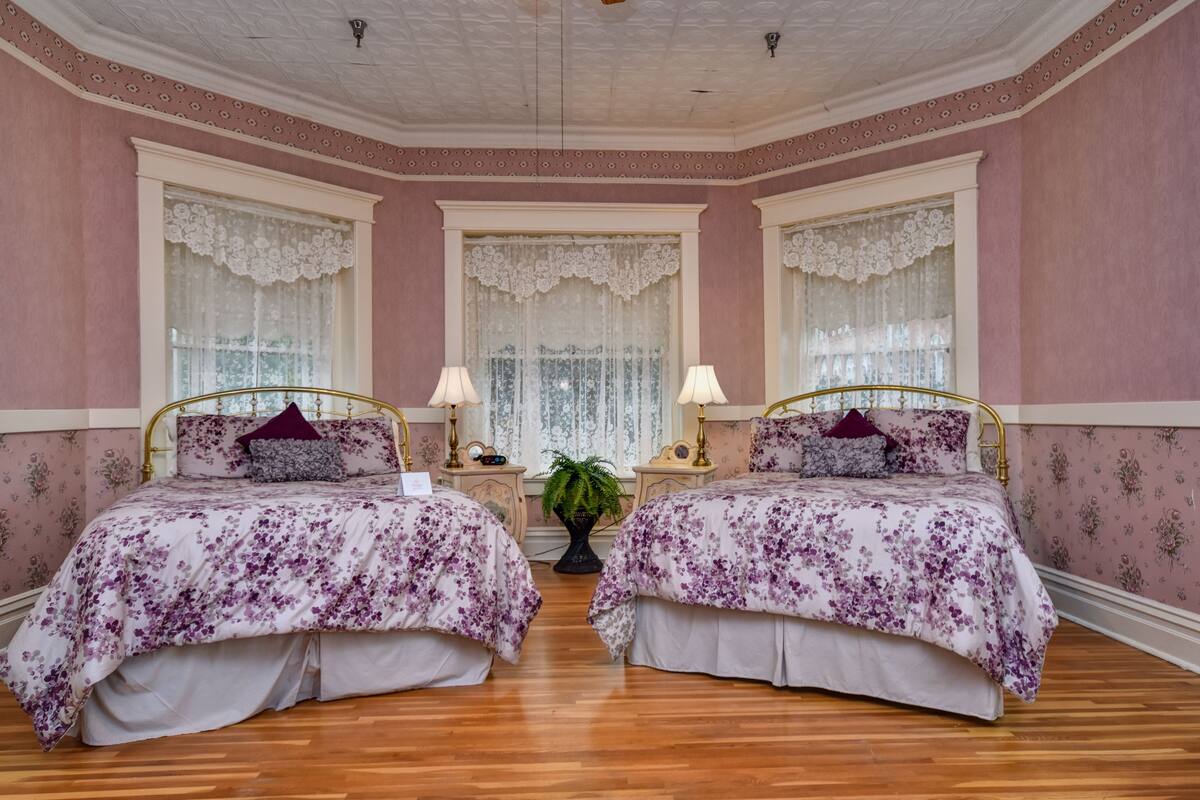 A room with two beds, each with a floral bedspread, a nightstand with a lamp, and a potted plant between the beds.