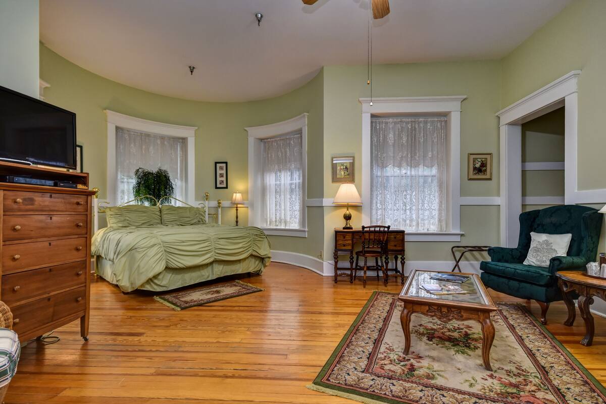A spacious living room with a large bed, a television, a green armchair, and a coffee table.