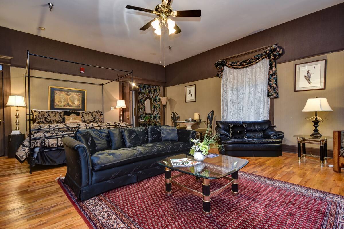 A spacious living room with a large black leather sofa, a glass coffee table, and a red patterned rug. There is a ceiling fan, a bed with a patterned headboard, and a large window with curtains.
