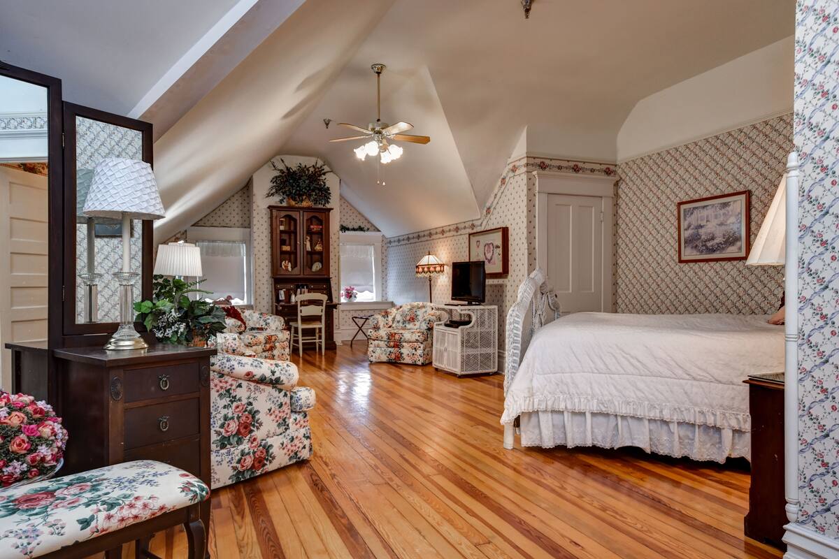 A spacious attic bedroom with a sloped ceiling, wooden flooring, a large bed, a desk, a chair, and a television.