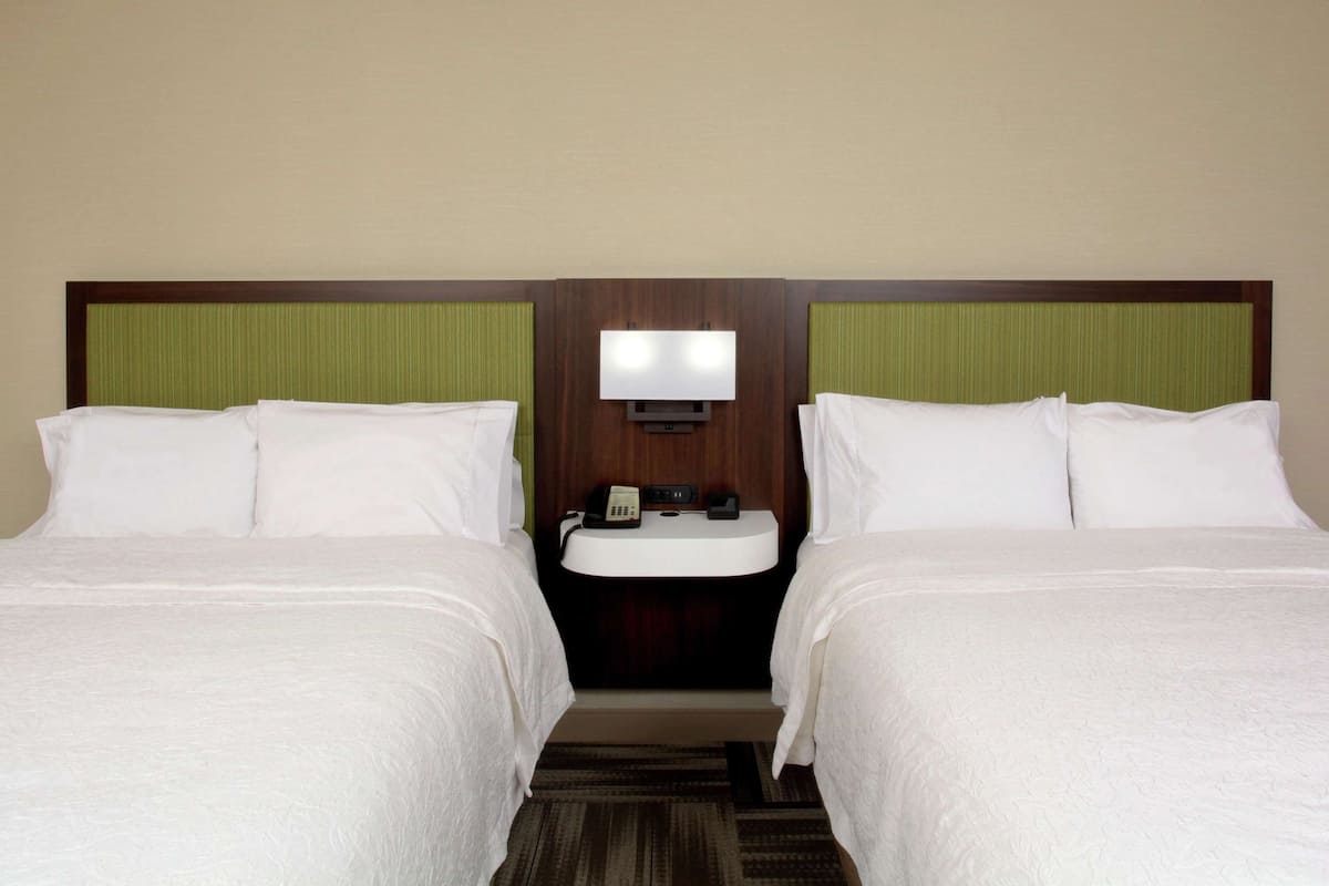 A hotel room with two beds, a bedside table with a phone and a lamp, and a small sink.