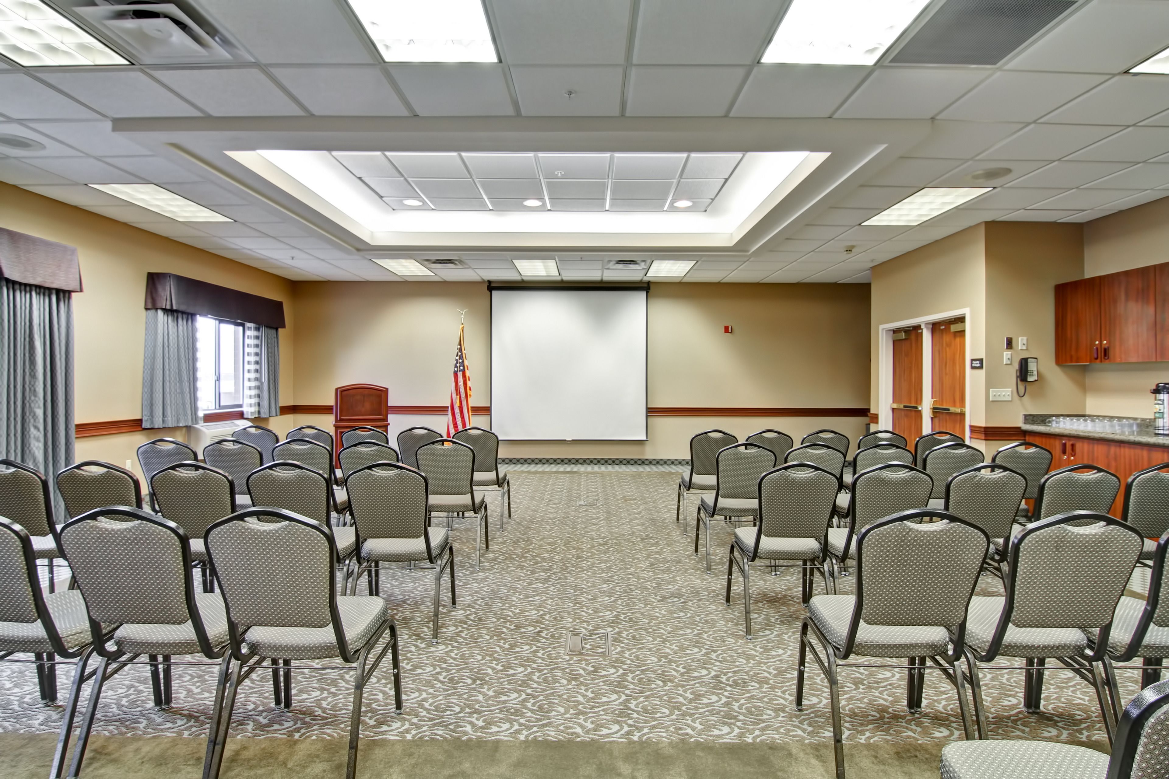 A conference room with rows of chairs, a projector screen, an American flag, and a carpeted floor.