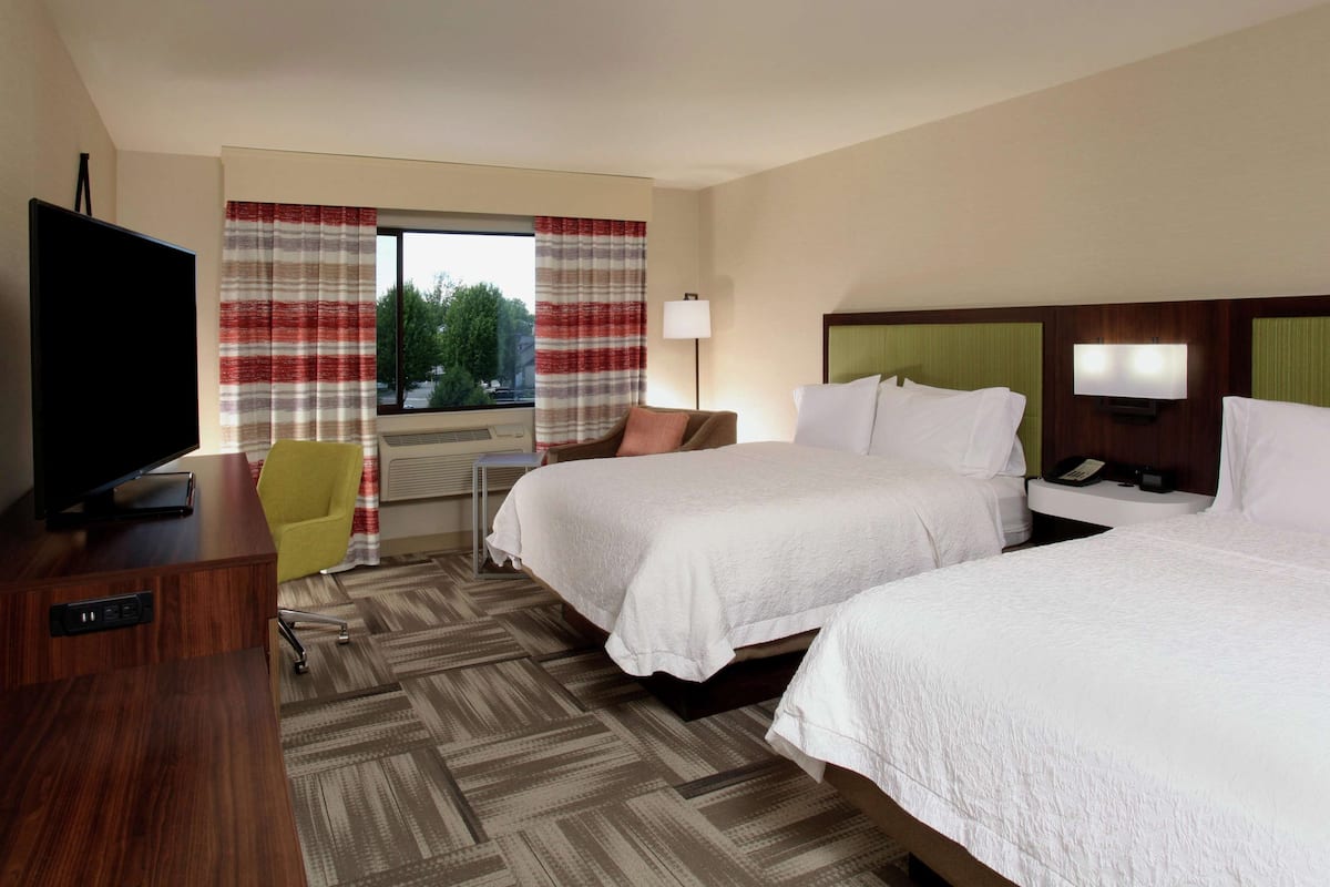 A hotel room with two beds, a TV, a desk, a chair, and a window with curtains.