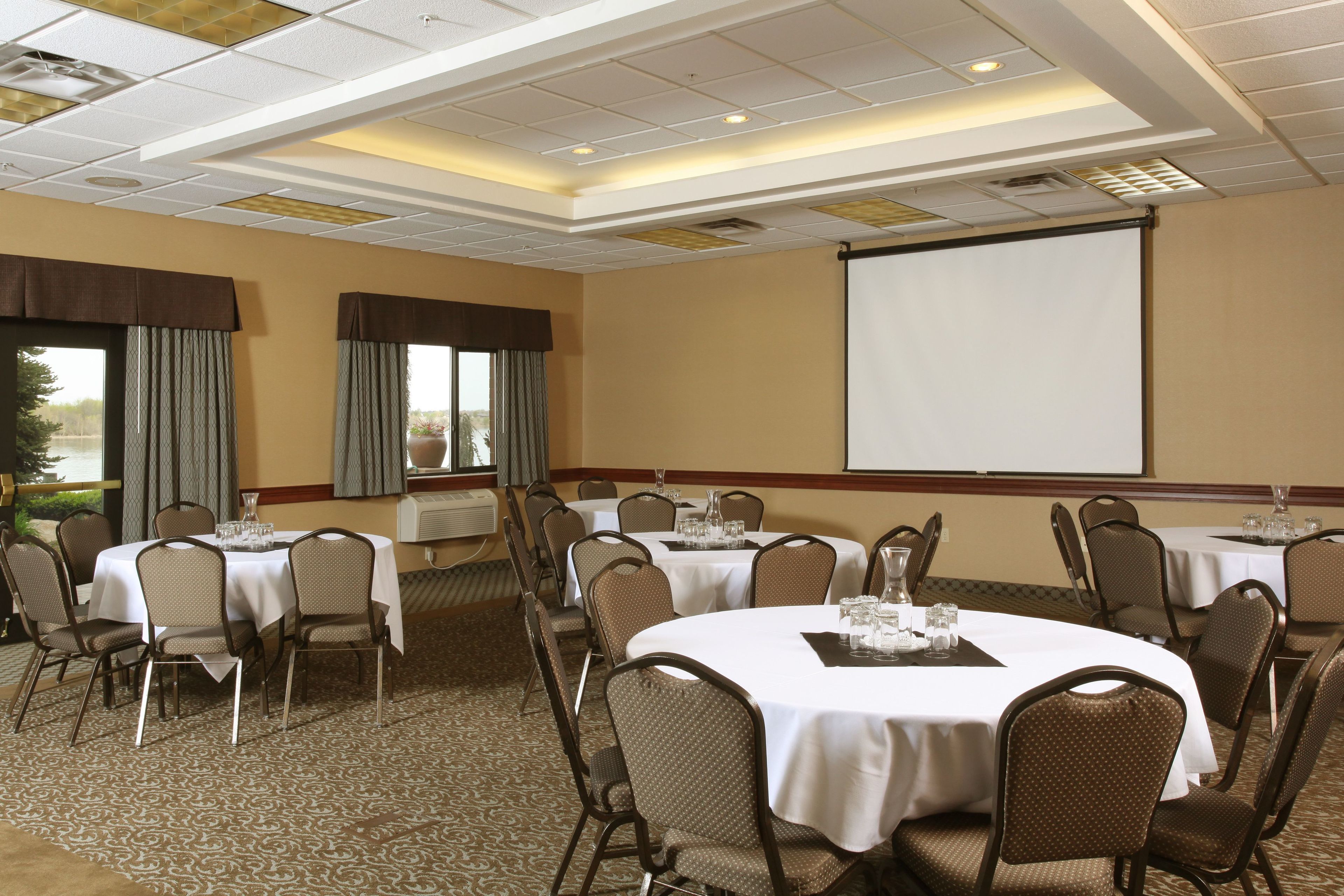 A conference room with multiple round tables, each with a white tablecloth and brown chairs. There is a large projection screen on the wall and a window with curtains.