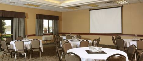 A conference room with multiple round tables, each with a white tablecloth and brown chairs. There is a large projection screen on the wall and a window with curtains.