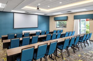 Un salón de conferencias con una larga mesa, sillas azules, una pantalla de proyector y ventanas con cortinas.