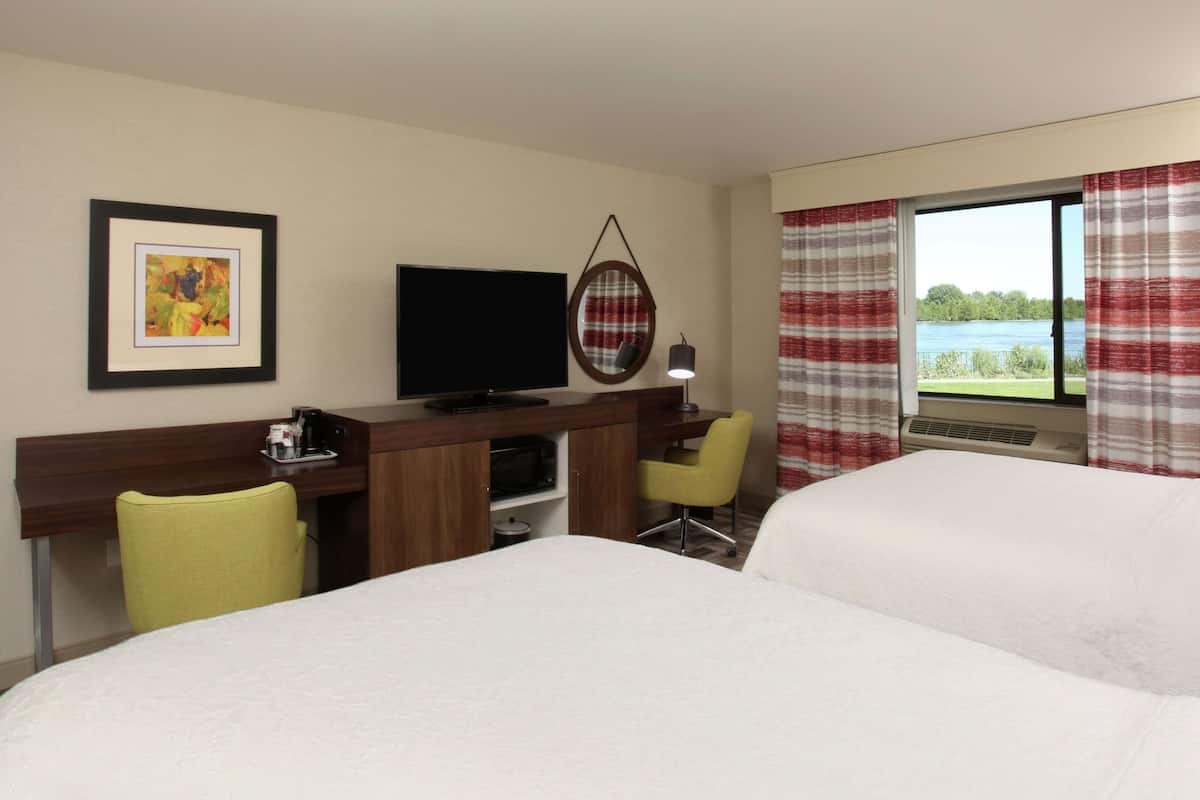 A hotel room with two beds, a desk with a television, a chair, and a view of a lake through the window.