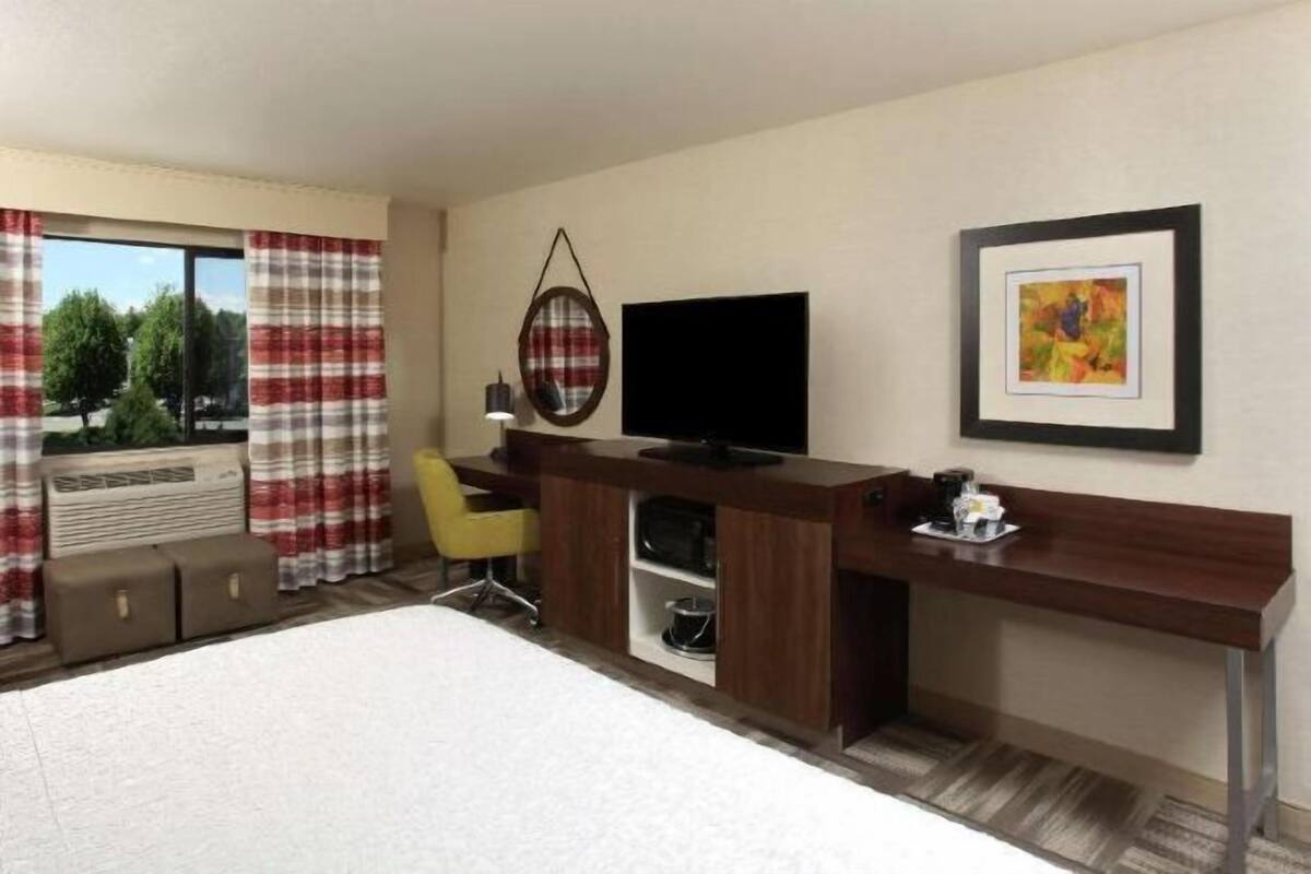 A hotel room with a bed, a desk with a flat-screen TV, a chair, a small table with a cup, and a window with curtains.