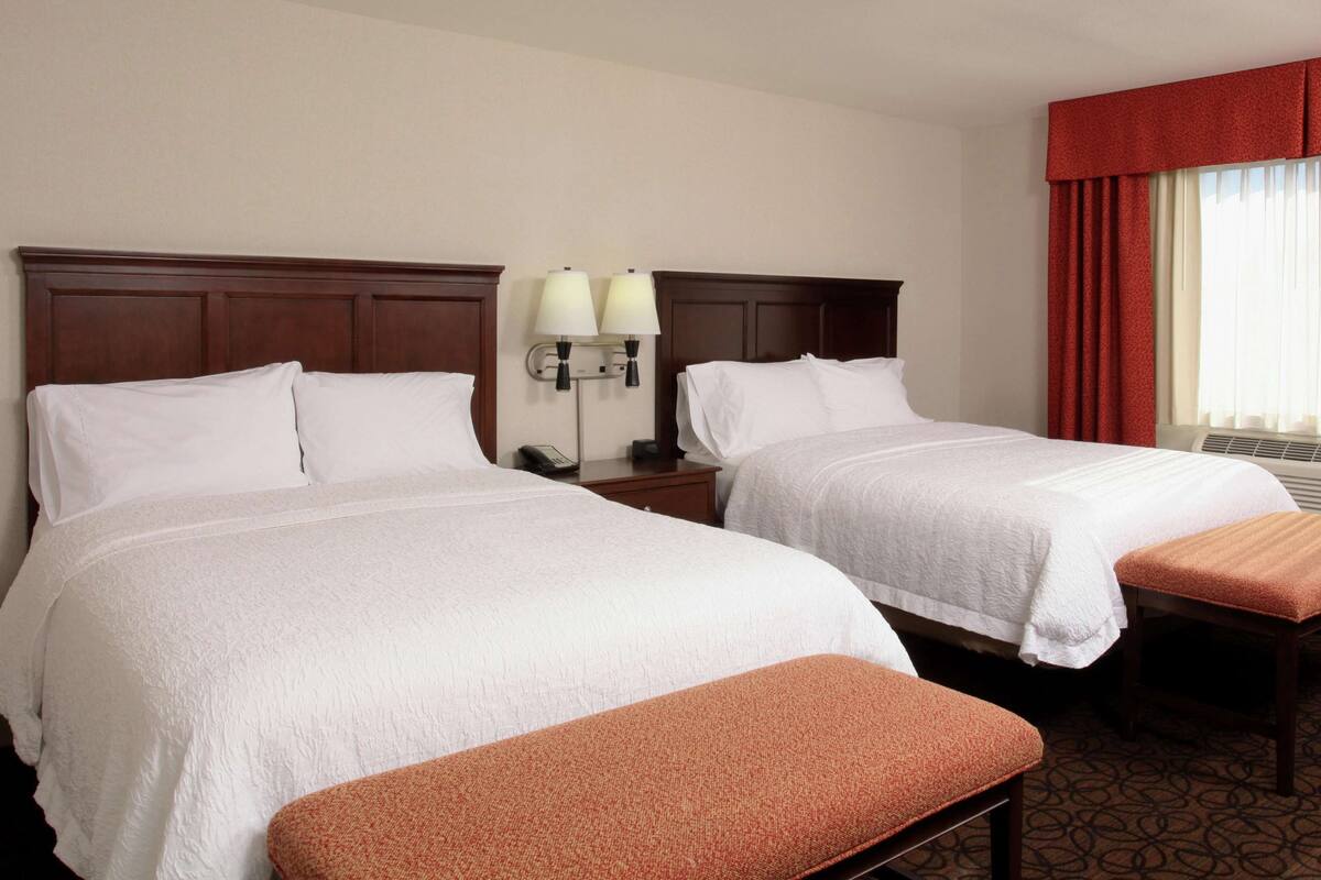 Two single beds with white linens, a wooden headboard, a nightstand with a lamp, and a red curtain.