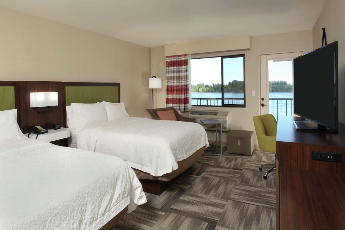A hotel room with two beds, a desk, a chair, a TV, and a view of a lake.