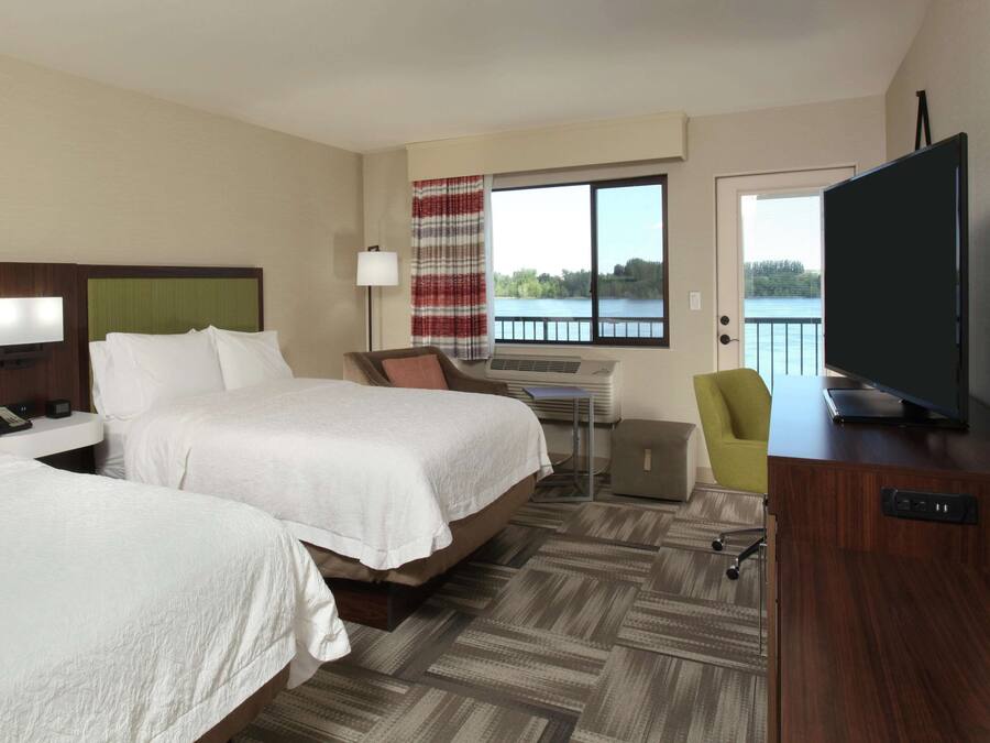 A hotel room with two beds, a desk, a chair, a TV, and a view of a lake.