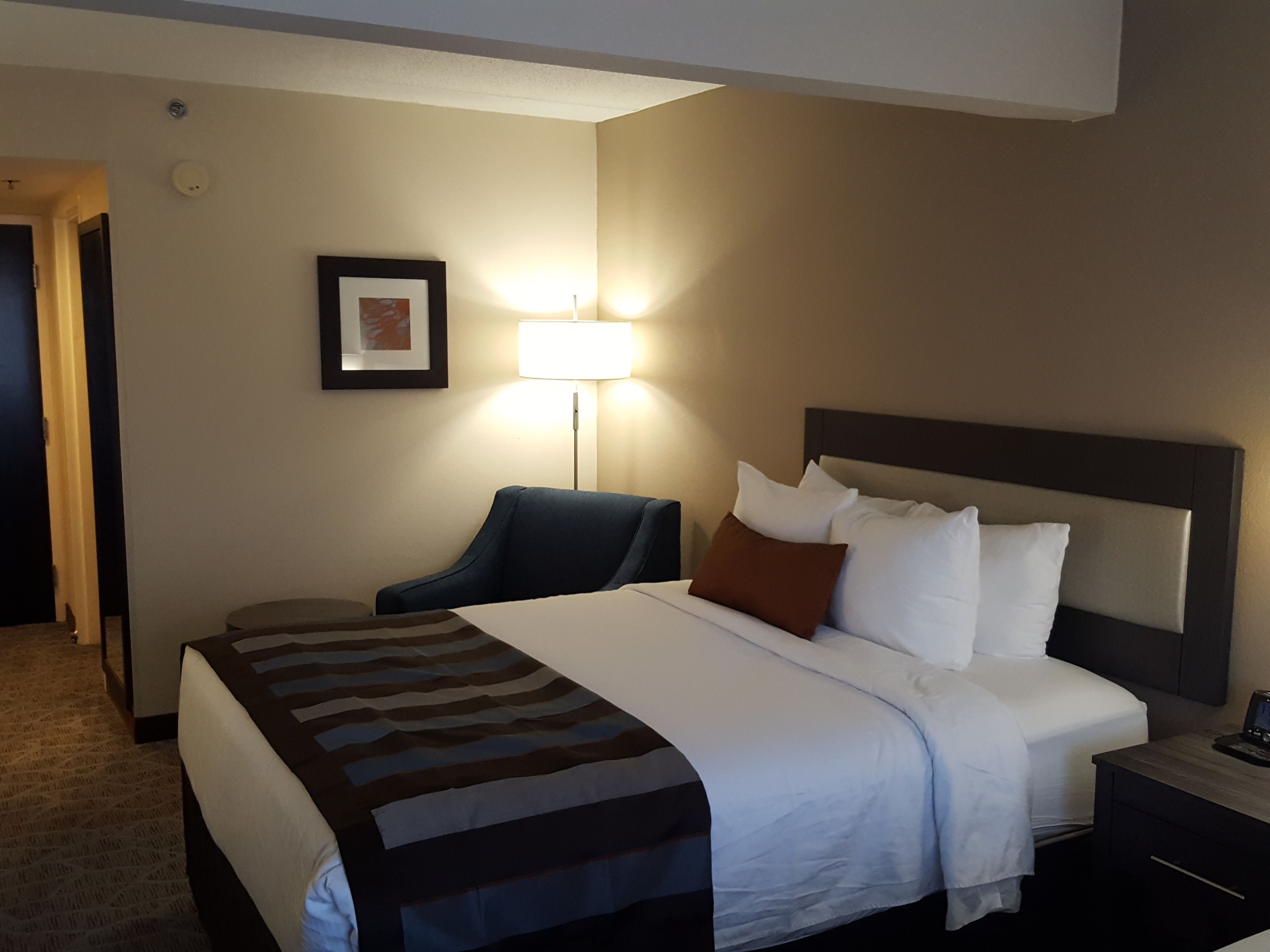 A hotel room with a large bed, a bedside table, a chair, a lamp, and a framed picture on the wall.