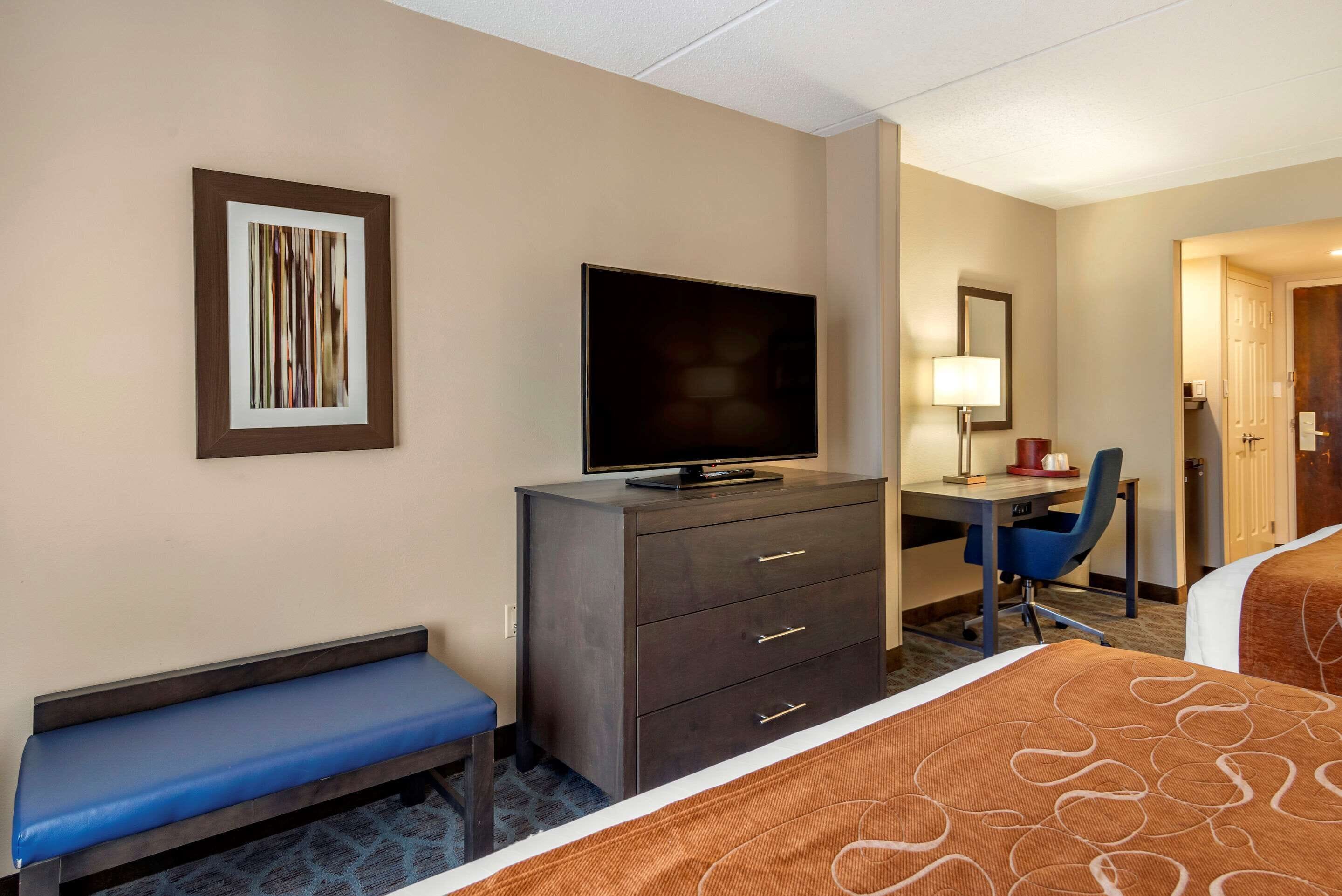 Quarto de hotel com uma cama, uma escrivaninha com cadeira, televisão, um banco e um quadro na parede.