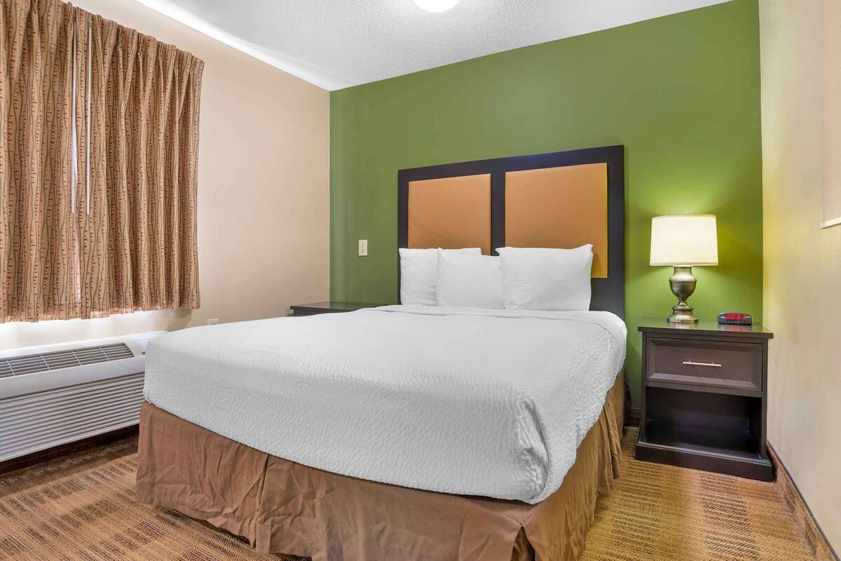 A hotel room with a large bed, a nightstand with a lamp, a window with curtains, and a wall heater.