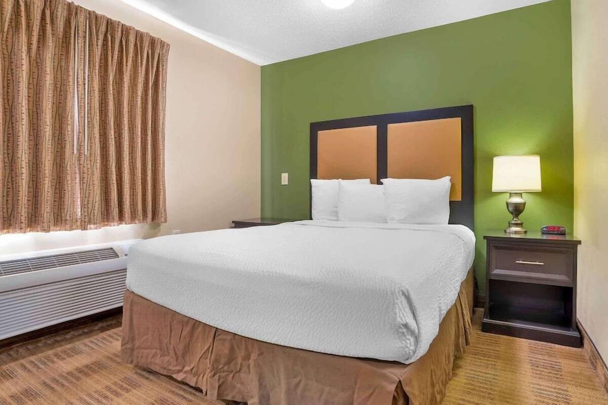 A hotel room with a large bed, a nightstand with a lamp, a window with curtains, and a wall heater.
