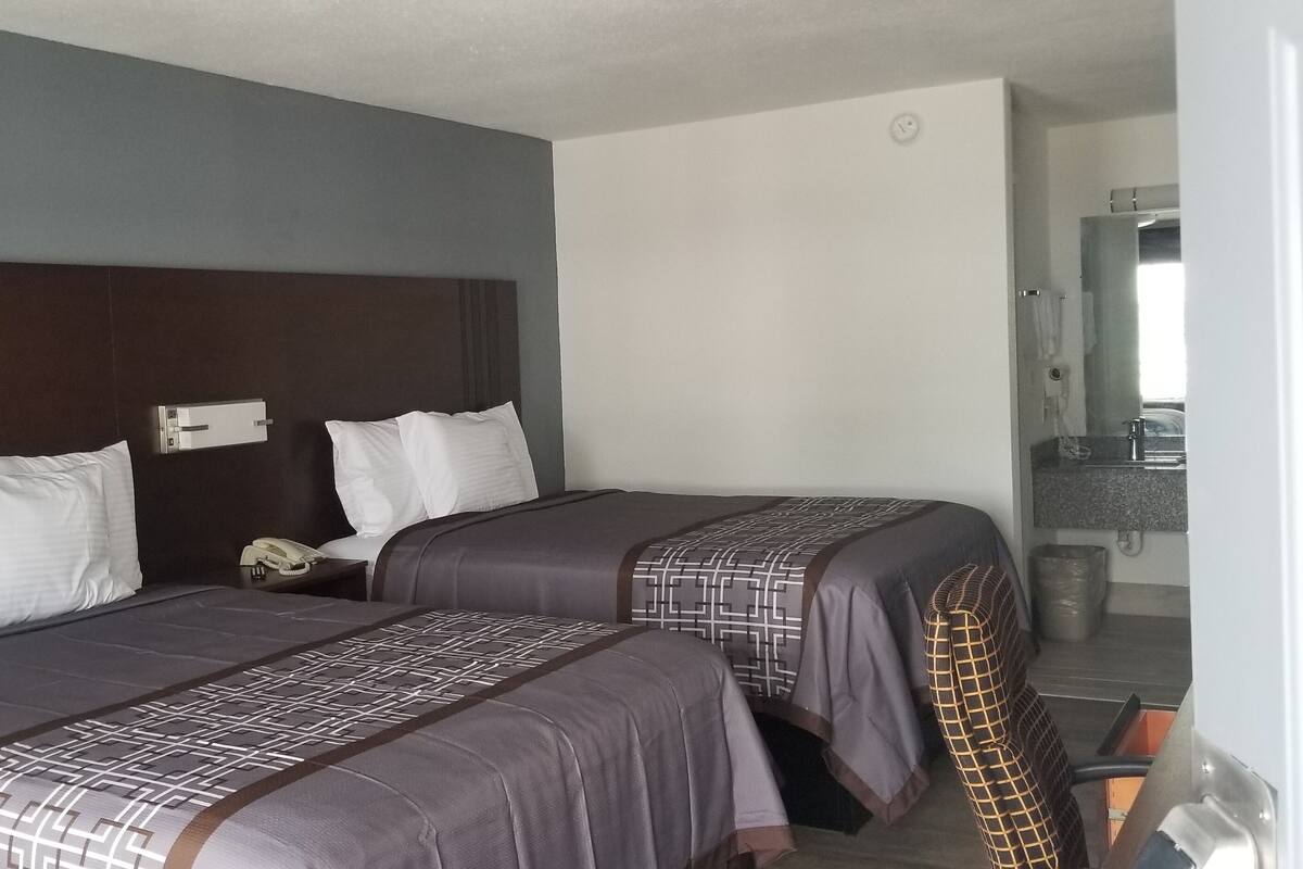A hotel room with two beds, each with a patterned bedspread and white pillows. There is a bedside table with a telephone and a wall-mounted light switch. The room has a wooden headboard and a plain wall.