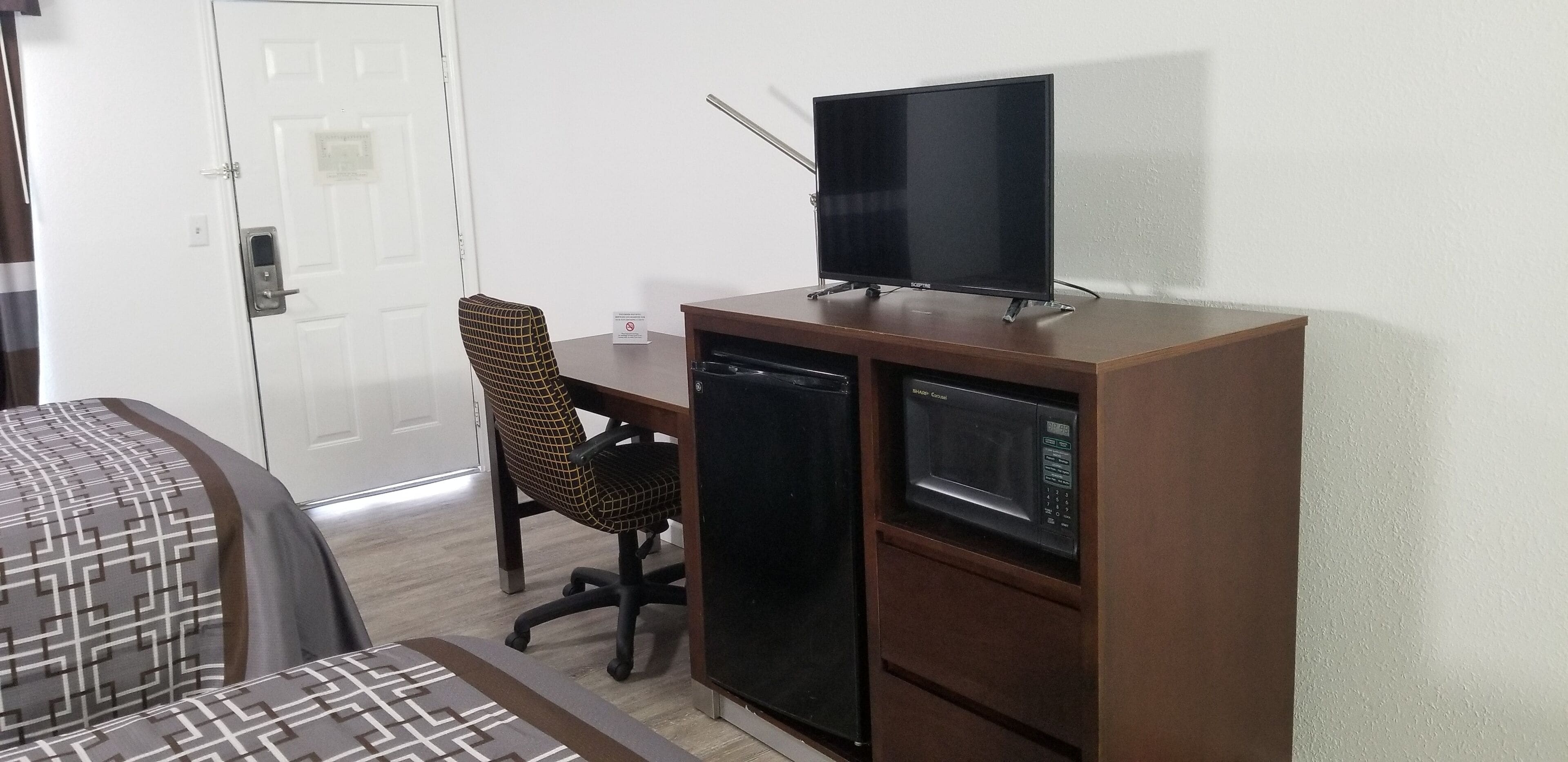 Quarto compacto com cama, escrivaninha com televisão, micro-ondas e uma cadeira.