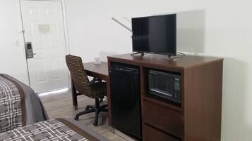Quarto compacto com cama, escrivaninha com televisão, micro-ondas e uma cadeira.