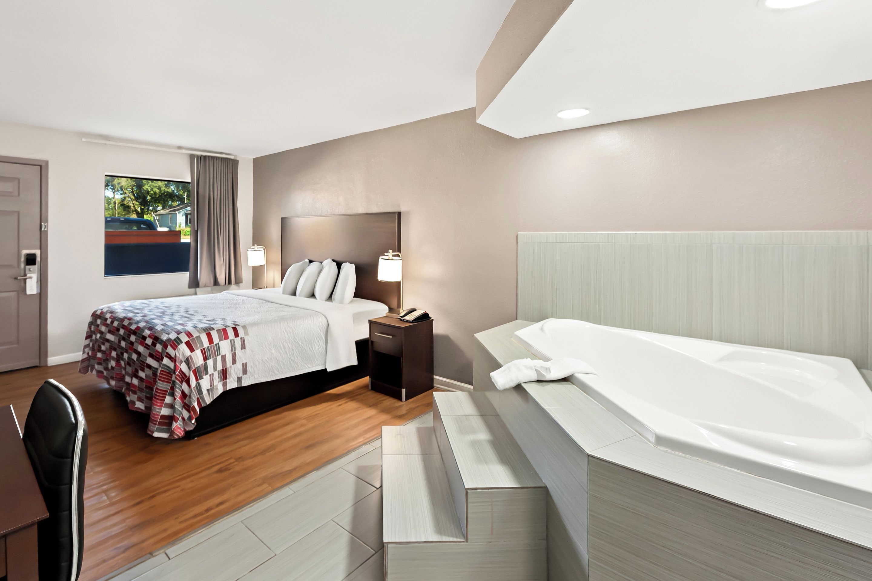 A modern hotel room with a large bed, a separate sitting area, a bathtub, and a view of the outdoors.