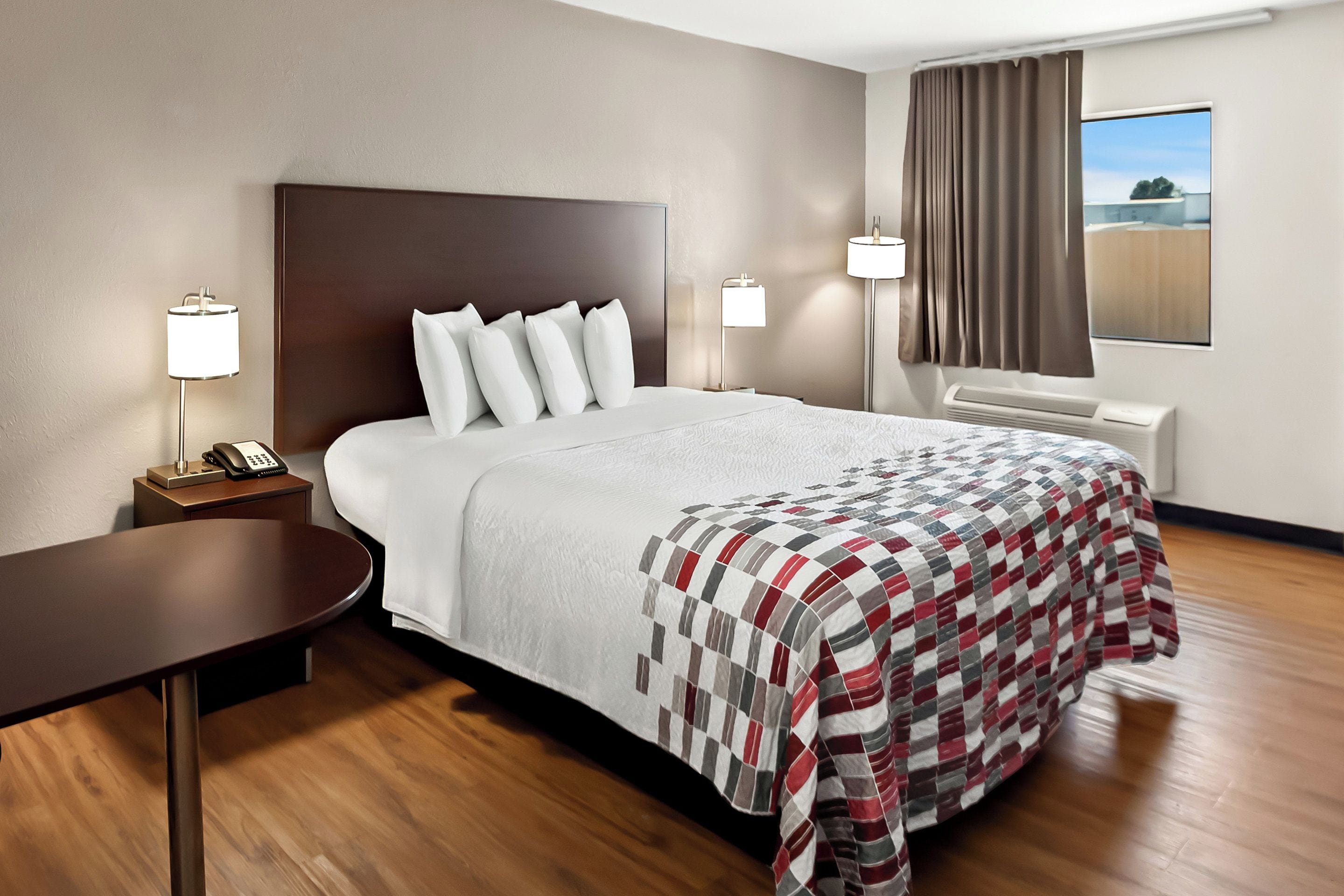 A hotel room with a large bed, a bedside table with a lamp, a window with curtains, and a view of the outside.