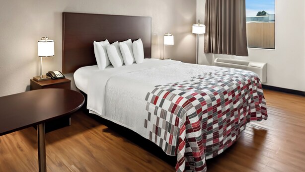 A hotel room with a large bed, a bedside table with a lamp, a window with curtains, and a view of the outside.
