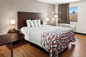 A hotel room with a large bed, a bedside table with a lamp, a window with curtains, and a view of the outside. - Red Roof Inn Muscle Shoals (Muscle Shoals)