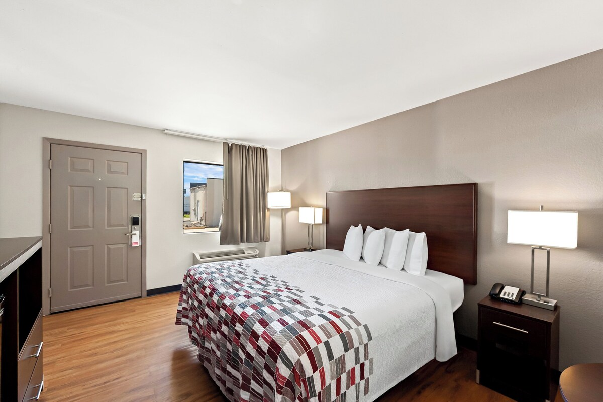 A hotel room with a large bed, a desk, a chair, a lamp, and a view of the outside.